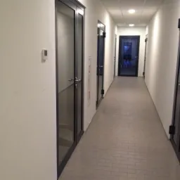 Długi korytarz biurowy z białymi ścianami i szarymi płytkami podłogowymi. Czarne, szklane drzwi po obu stronach korytarza. Oświetlenie sufitowe.