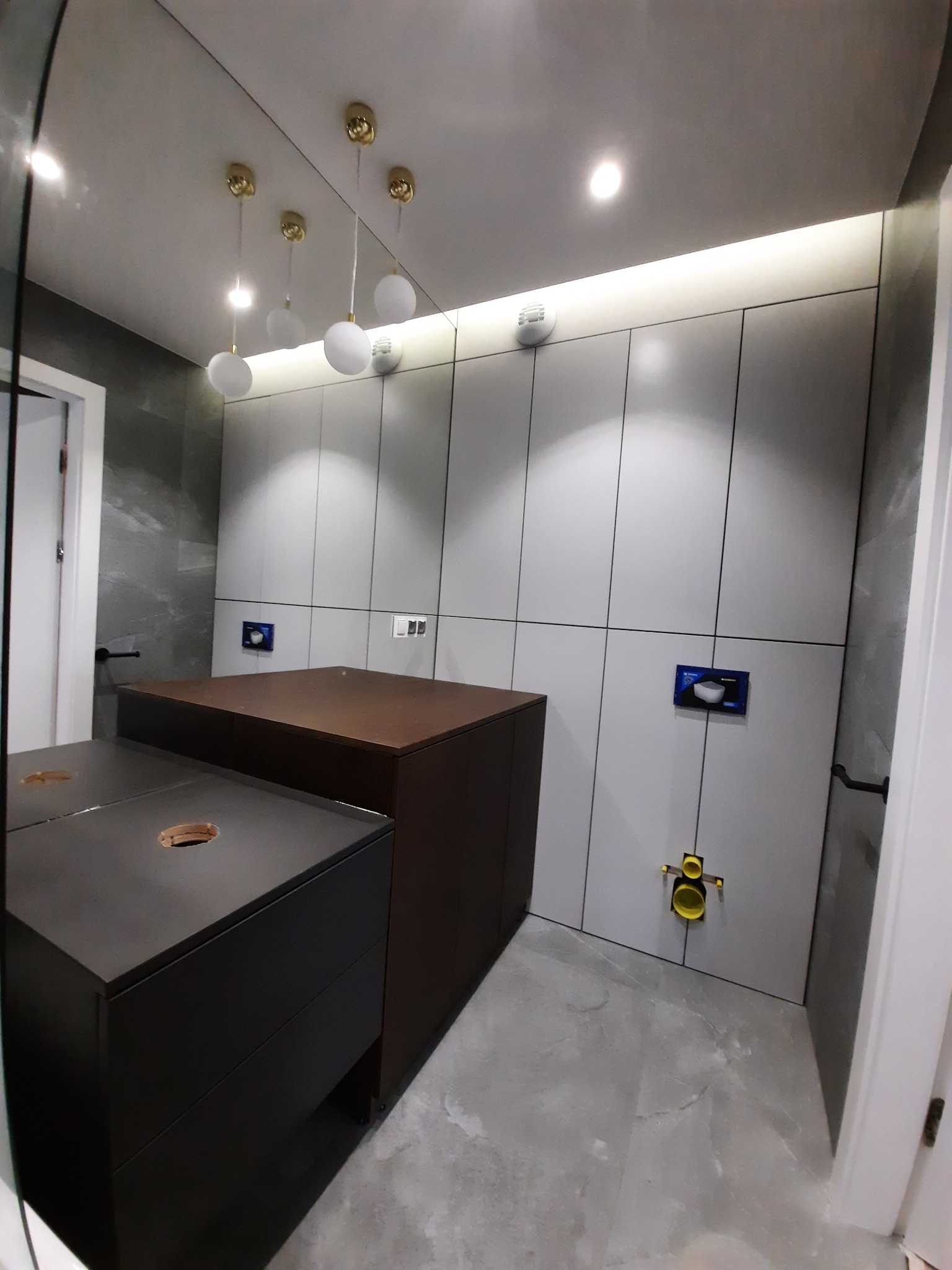 Łazienka z szarymi szafkami na wymiar, drewnianym blatem i oświetleniem LED pod sufitem, widoczne przyłącza hydrauliczne.