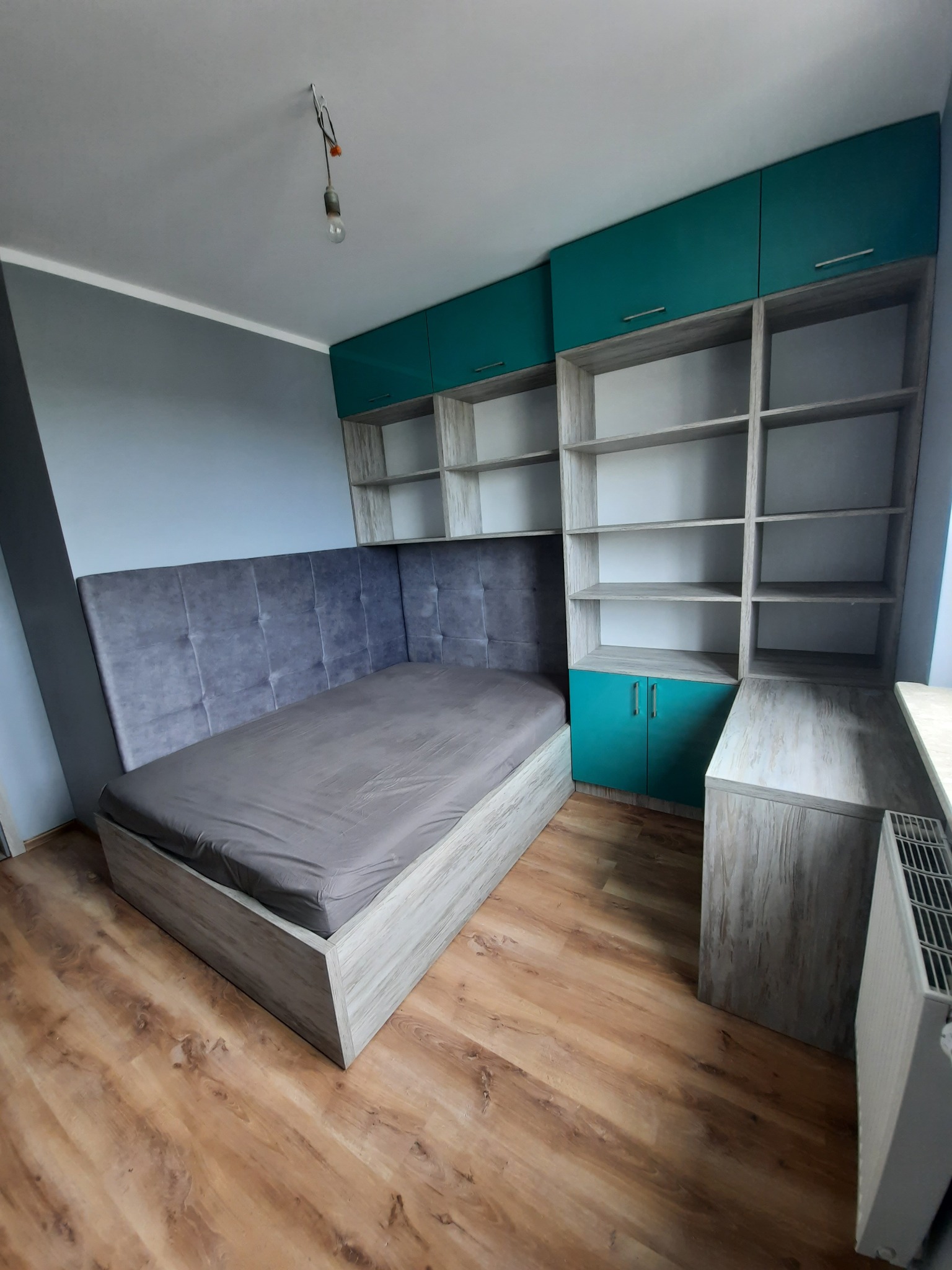 Szafa wnękowa zintegrowana z łóżkiem i regałem, fronty szafek w kolorze turkusowym, szare tapicerowane wezgłowie, surowa żarówka zwisająca z sufitu.