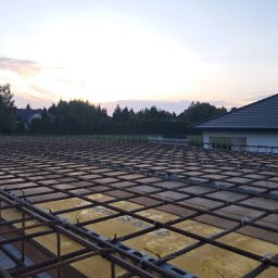 Paweł Borowski - Zbrojenie fundamentu domu jednorodzinnego, widoczne siatki stalowe i żółte szalunki, w tle zachodzące słońce i zieleń drzew.