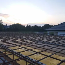 Zbrojenie fundamentu domu jednorodzinnego, widoczne siatki stalowe i żółte szalunki, w tle zachodzące słońce i zieleń drzew.