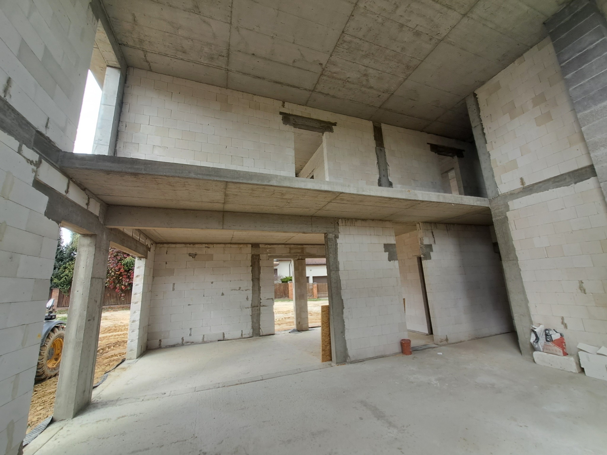 Wnętrze dwukondygnacyjnego domu w trakcie budowy, widoczne ściany z białych bloczków, betonowe stropy, na zewnątrz widoczny traktor z żółtymi kołami i fragment zabudowy jednorodzinnej.