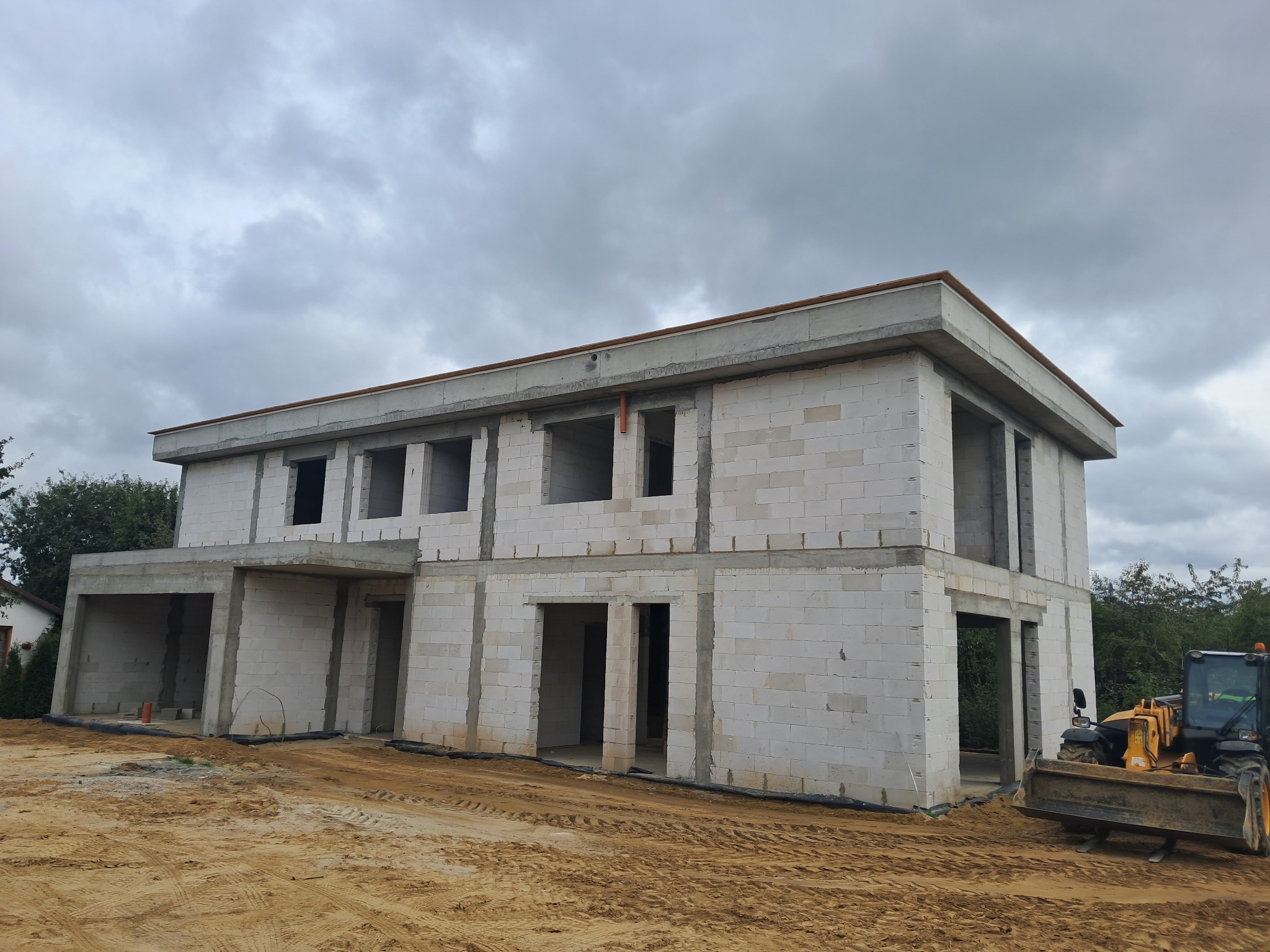 Dwukondygnacyjny dom w stanie surowym otwartym, zbudowany z białych bloczków, z widoczną żółtą maszyną budowlaną na piaszczystym terenie, pod pochmurnym niebem.