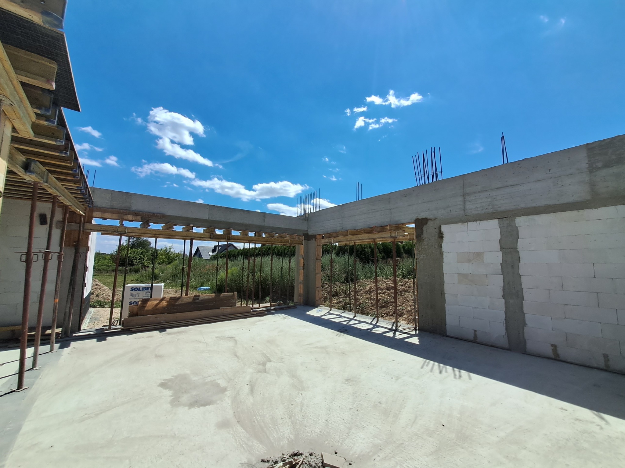 Wnętrze surowego domu z betonu komórkowego w trakcie budowy, widoczne betonowe nadproża z wystającymi prętami zbrojeniowymi, drewniane szalunki oraz podpory stalowe, w tle błękitne niebo z chmurami.