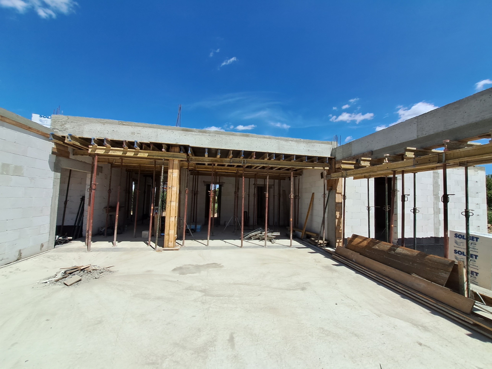 Surowy stan domu w trakcie budowy: widoczne białe ściany z betonu komórkowego, drewniane szalunki podtrzymujące strop, metalowe stemple budowlane, błękitne niebo w tle.