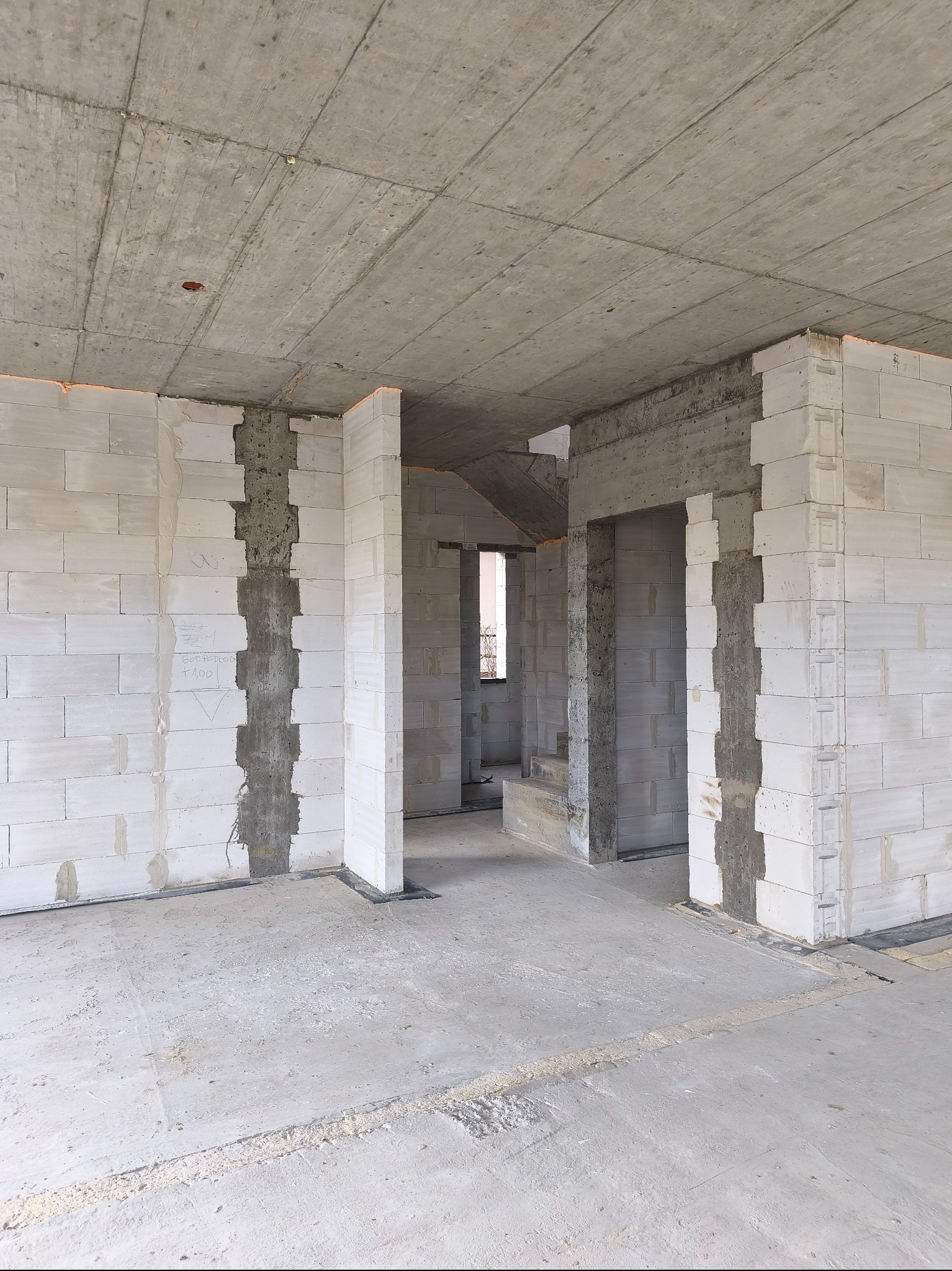 Wnętrze domu w trakcie budowy: ściany z białych bloczków, surowy betonowy strop, widoczne otwory drzwiowe i okienne. Etap murowania ścian.