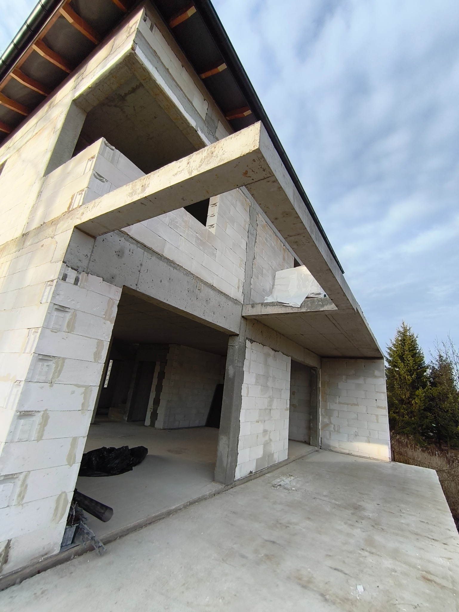 Surowy stan domu z betonu komórkowego w trakcie budowy, widok z dołu, ukazujący konstrukcję ścian, stropów i otworów okiennych na tle pochmurnego nieba.