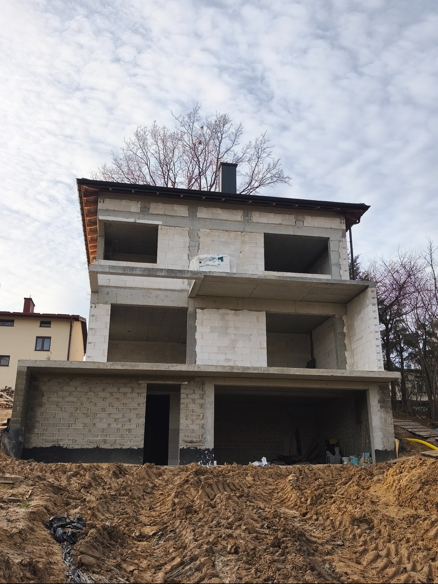 Surowy, dwupiętrowy dom z betonu komórkowego w trakcie budowy, z widocznymi otworami okiennymi i drzwiami, na tle pochmurnego nieba i ziemi z wyraźnymi śladami opon.