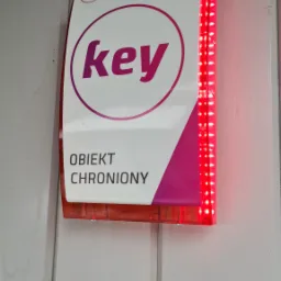 Zewnętrzna sygnalizacja alarmowa z napisem 'Obiekt Chroniony' i pulsującymi czerwonymi diodami LED, zamontowana na białej drewnianej elewacji.