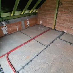 Instalacja elektryczna w surowym stanie: czerwone i szare kable ułożone na wylewce podłogowej w pomieszczeniu z ceglanymi ścianami i drewnianą konstrukcją dachu.