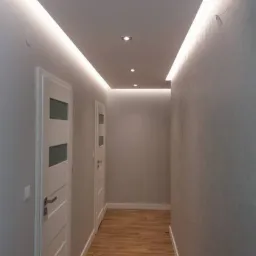 Korytarz w mieszkaniu po remoncie z podwieszanym sufitem z oświetleniem LED i drewnopodobną podłogą z płytek, widoczne białe drzwi z przeszkleniami.