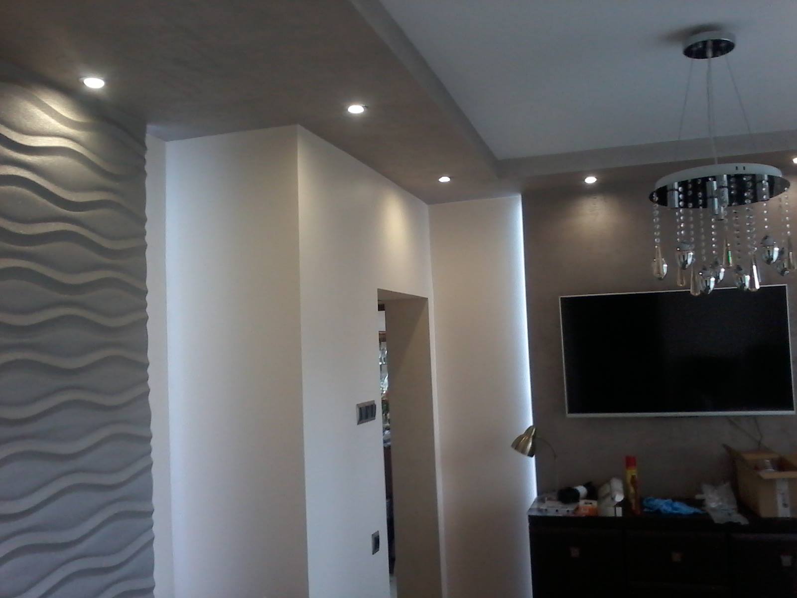 Wnętrze salonu po remoncie: dekoracyjna ściana z panelami 3D o falistym wzorze, sufit podwieszany z oświetleniem punktowym, nowoczesny żyrandol z kryształkami nad telewizorem.