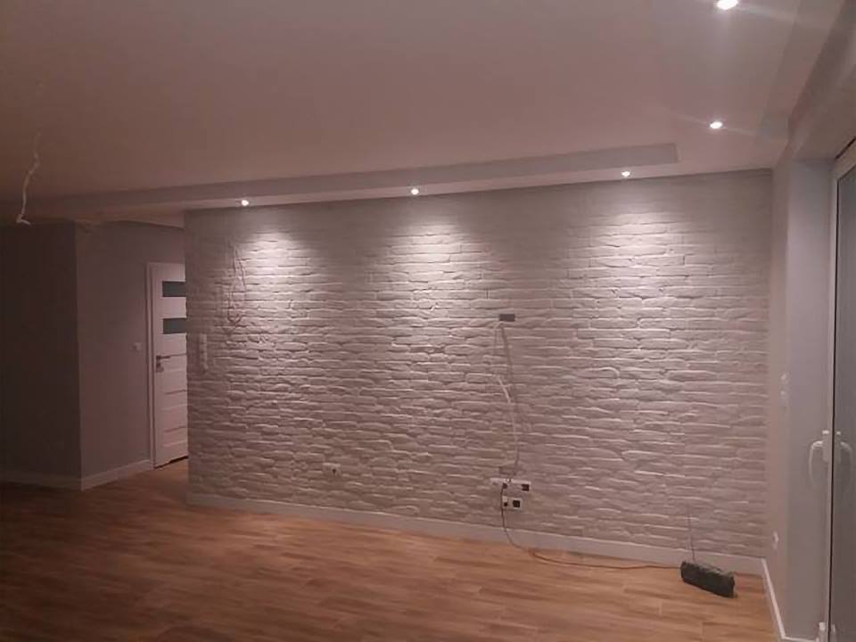 Ściana wyłożona białym kamieniem dekoracyjnym, oświetlona punktowymi lampami sufitowymi, z widocznymi gniazdkami elektrycznymi i częściowo odsłoniętymi kablami.