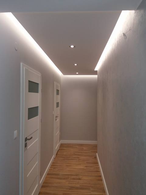 Korytarz w mieszkaniu po remoncie z podwieszanym sufitem z oświetleniem LED i drewnopodobną podłogą z płytek, widoczne białe drzwi z przeszkleniami.