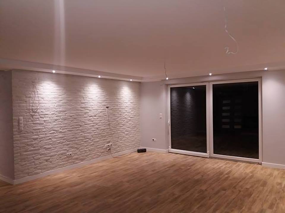 Wykończone wnętrze z dekoracyjną ścianą z białej cegły, drewnianą podłogą i przesuwnymi drzwiami balkonowymi, widoczne punkty świetlne w suficie.