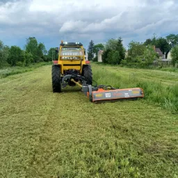 Żółty traktor z kosiarką rotacyjną na polu świeżo skoszonej trawy, widok z przodu na tle zabudowań wiejskich pod pochmurnym niebem.