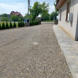Podjazd z szarego kruszywa otoczony niskimi tujami i chodnikiem z szarej kostki brukowej prowadzący do domu, widok na zadbany ogród i inne budynki w tle.