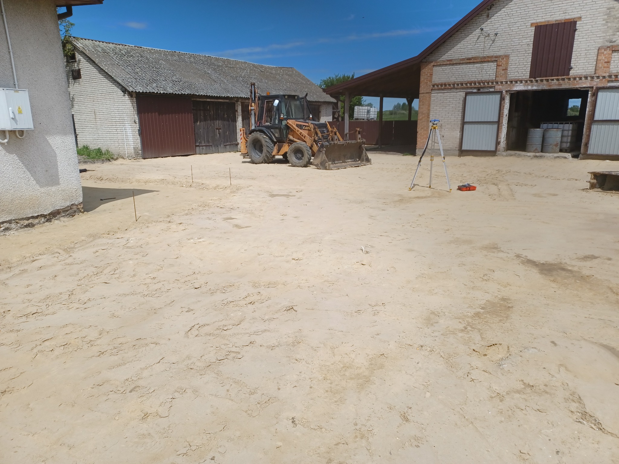 Żółta koparko-ładowarka na piaszczystym terenie z budynkami gospodarczymi w tle, widoczny niwelator optyczny, teren przygotowany pod dalsze prace.