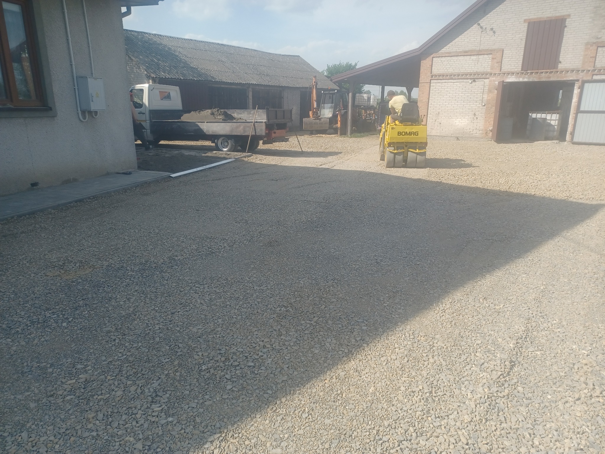 Żółty walec drogowy Bomag ubija szarą nawierzchnię z kruszywa na placu budowy, w tle budynki gospodarcze i ciężarówka z ładunkiem ziemi.