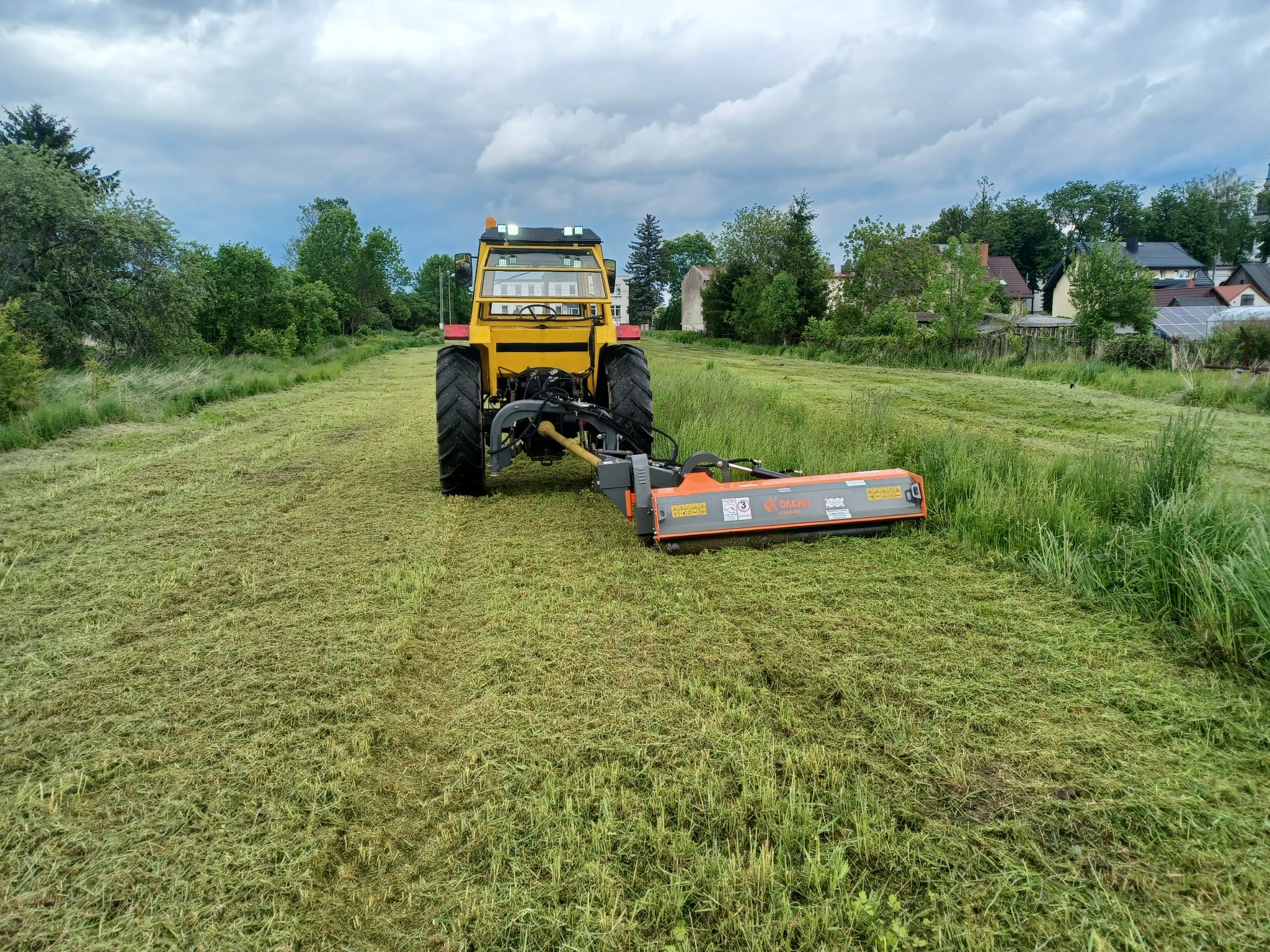Żółty traktor z kosiarką rotacyjną na polu świeżo skoszonej trawy, widok z przodu na tle zabudowań wiejskich pod pochmurnym niebem.