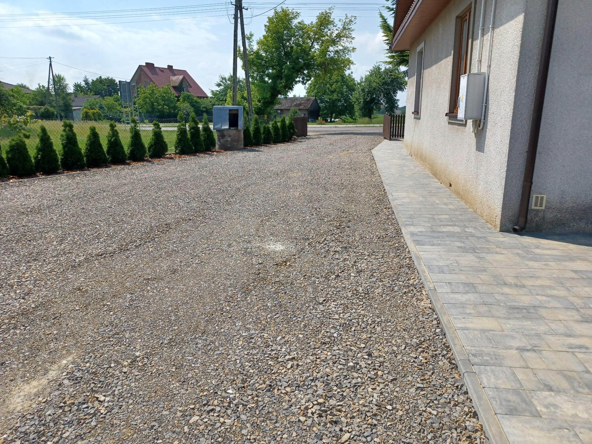 Podjazd z szarego kruszywa otoczony niskimi tujami i chodnikiem z szarej kostki brukowej prowadzący do domu, widok na zadbany ogród i inne budynki w tle.