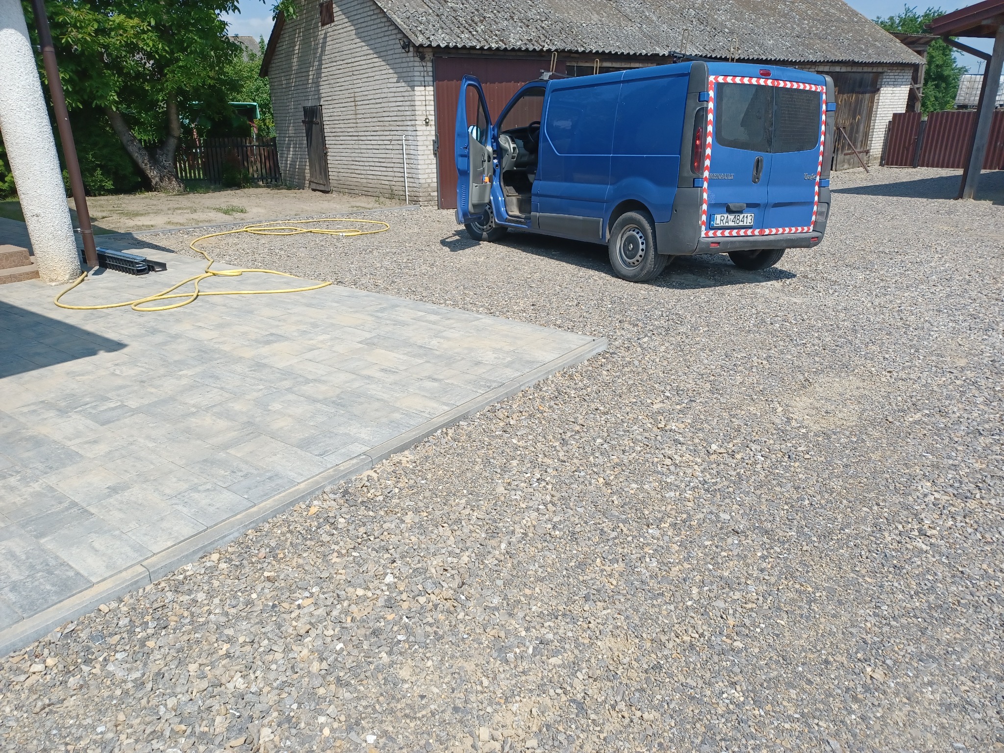 Ułożona szara kostka brukowa sąsiaduje z obszarem wysypanym drobnym żwirem, na którym stoi niebieski samochód dostawczy z otwartymi drzwiami. W tle widoczny budynek gospodarczy.