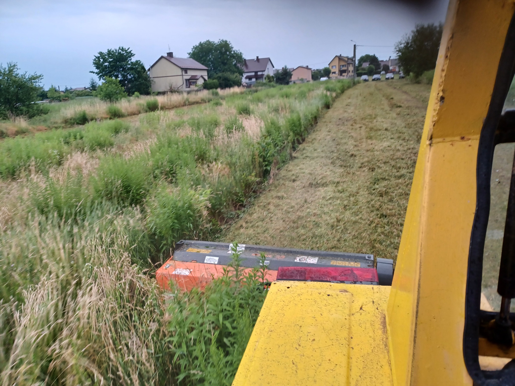 Widok z kabiny żółtej maszyny koszącej, na skoszoną trawę i niekoszone chwasty na poboczu drogi w oddali widać zabudowania.