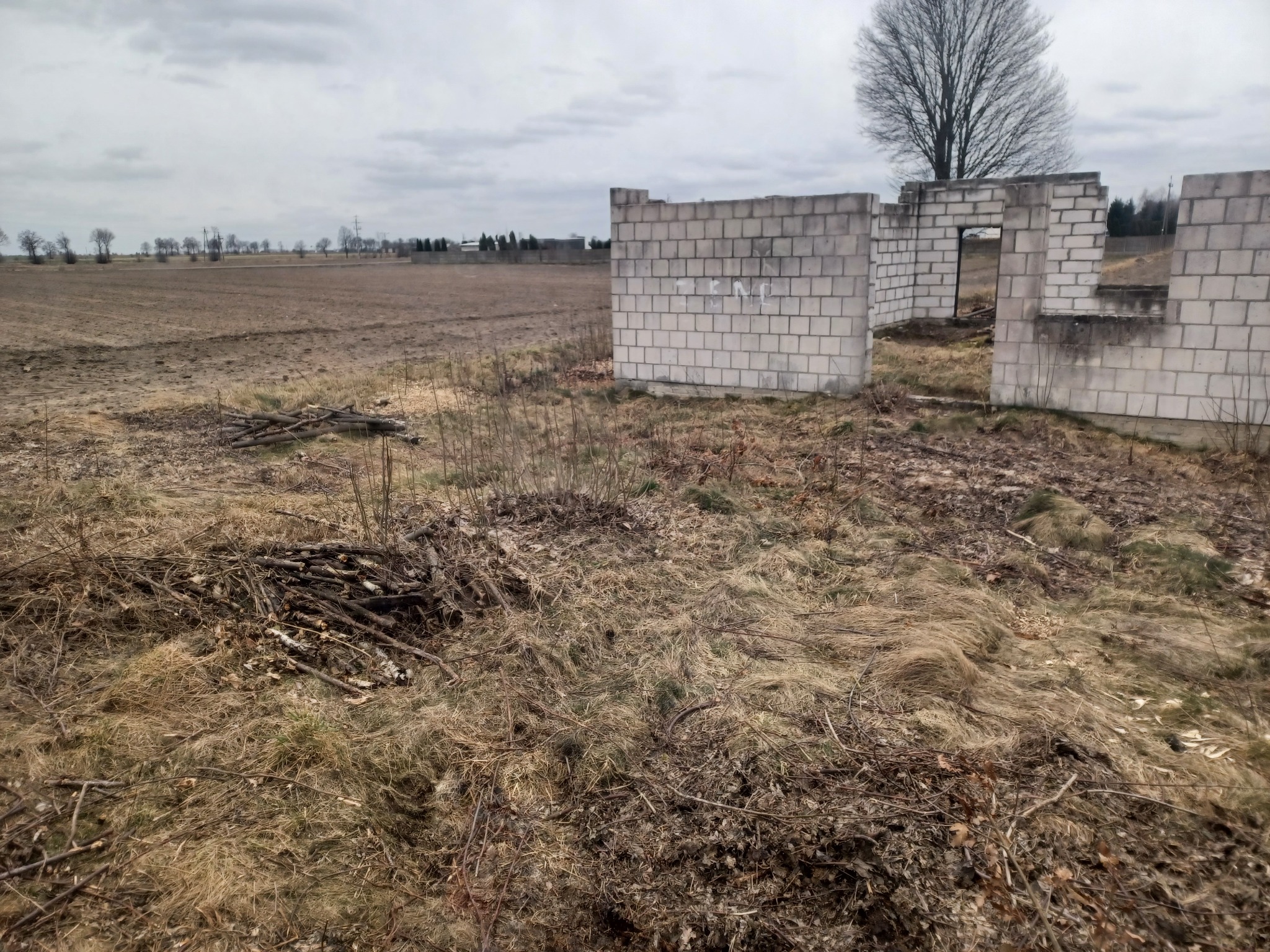 Surowy stan budowy: fragment muru z bloczków betonowych na zarośniętym terenie, w tle pole i pochmurne niebo.
