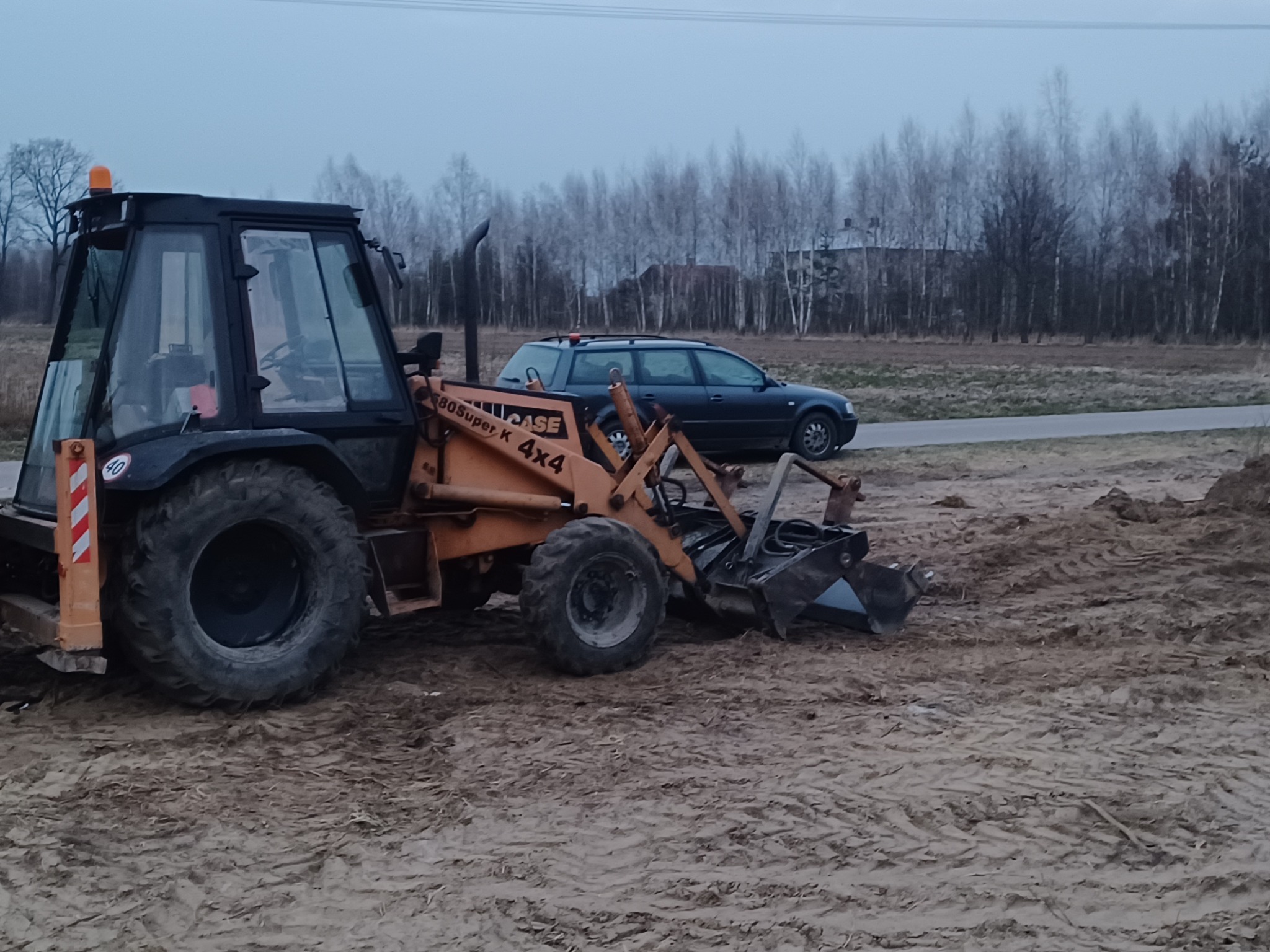 Pomarańczowo-czarna koparko-ładowarka Case 80 Super K 4x4 na placu budowy, z wykopaną ziemią, w tle zaparkowany samochód osobowy i rzadki las.