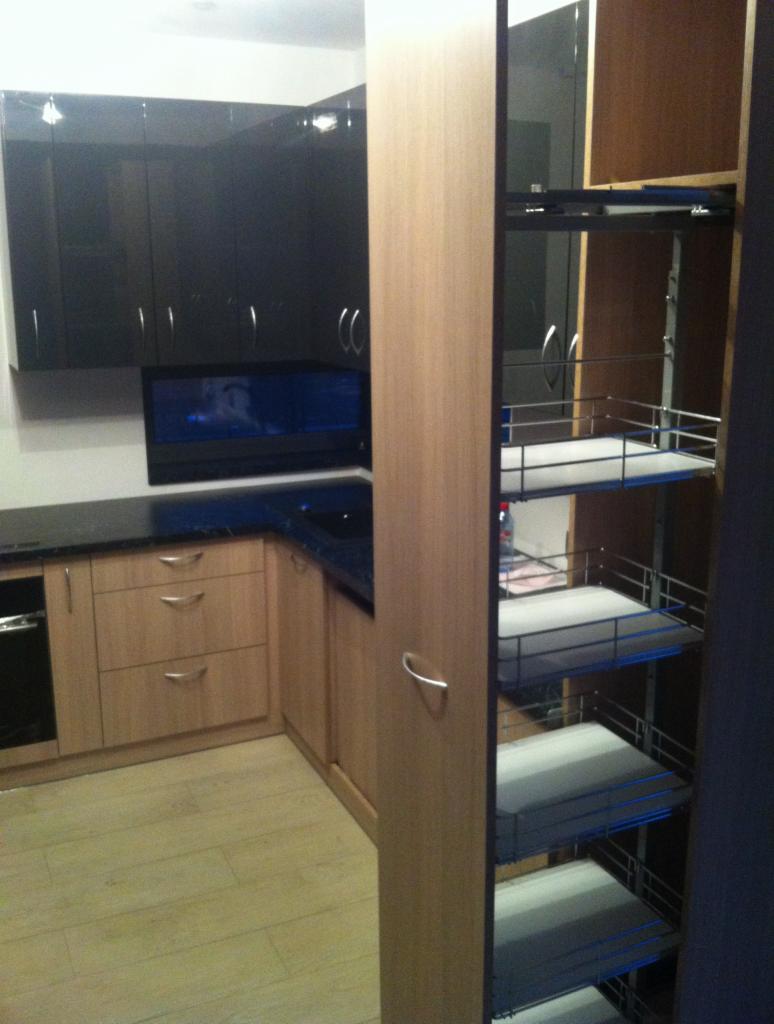 Nowoczesna kuchnia w odcieniach beżu i czerni z wysuwanym koszem cargo. Jasna podłoga i ciemny blat dodają kontrastu. Widoczne fronty szafek i metalowe akcenty.