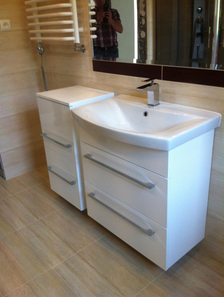 Biała umywalka z szafką i chromowaną baterią w łazience wykończonej beżowymi płytkami, odbicie fotografa w lustrze.