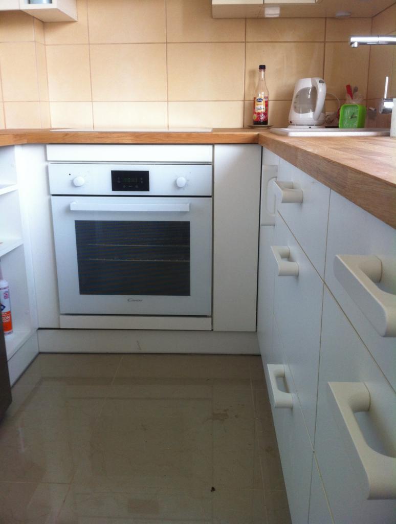 Biała zabudowa kuchenna z drewnianym blatem, beżowe płytki na ścianie. Widoczny piekarnik Candy, szafki z charakterystycznymi uchwytami i fragment podłogi z płytek.