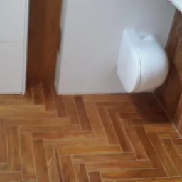 Ukończona łazienka z podłogą wyłożoną drewnopodobnymi płytkami w jodełkę, widoczna biała toaleta i fragment szafki.