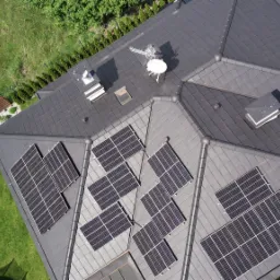 Widok z góry na dach domu z grafitową dachówką i panelami fotowoltaicznymi, antena satelitarna, komin wentylacyjny, zadbany trawnik i ozdobne nasadzenia.