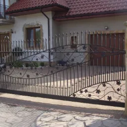 Elegancka, przesuwna brama kuta z motywem roślinnym, zamontowana przed domem z kamiennymi słupkami. Widoczny fragment podjazdu i elewacja budynku.