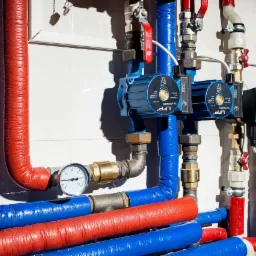 Instalacja centralnego ogrzewania z czerwono-niebieskimi rurami i dwoma pompami obiegowymi LFP, manometrem i zaworami na tle białych płytek.