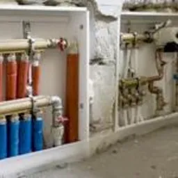 Rozdzielacz ogrzewania podłogowego w białej obudowie, z rurami w kolorze czerwonym i niebieskim, zamontowany w ścianie podczas prac wykończeniowych.