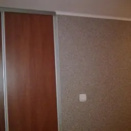 Wnętrze: ściana z dekoracyjnym tynkiem w odcieniach beżu, obok przesuwne drzwi szafy w kolorze drewna z srebrnymi ramami. Widoczny włącznik światła i fragment drzwi.