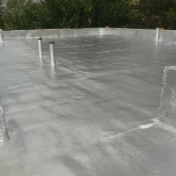 Szara, błyszcząca izolacja natryskowa na płaskim dachu z widocznymi kominkami wentylacyjnymi i fragmentem ceglanego komina.