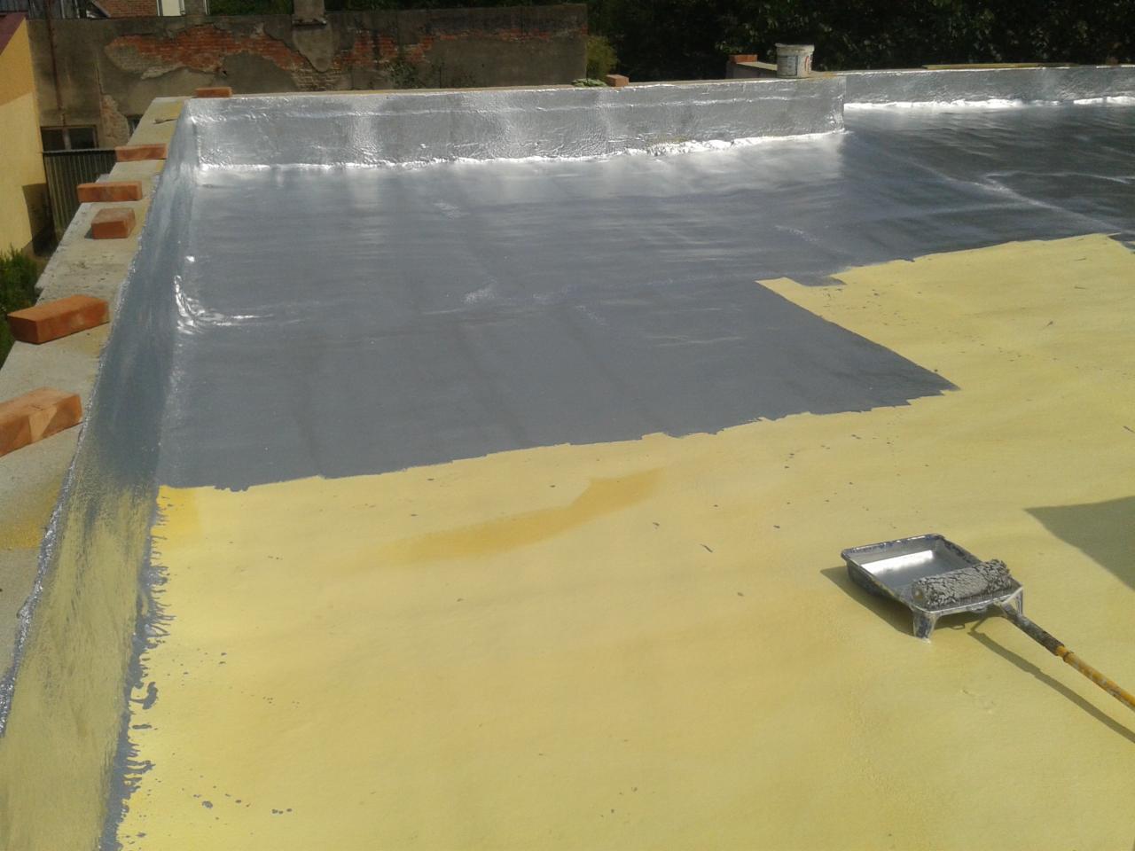 Szary płynny materiał izolacyjny w trakcie aplikacji na żółtą powierzchnię dachu płaskiego, widoczny wałek malarski i cegły na krawędzi dachu.