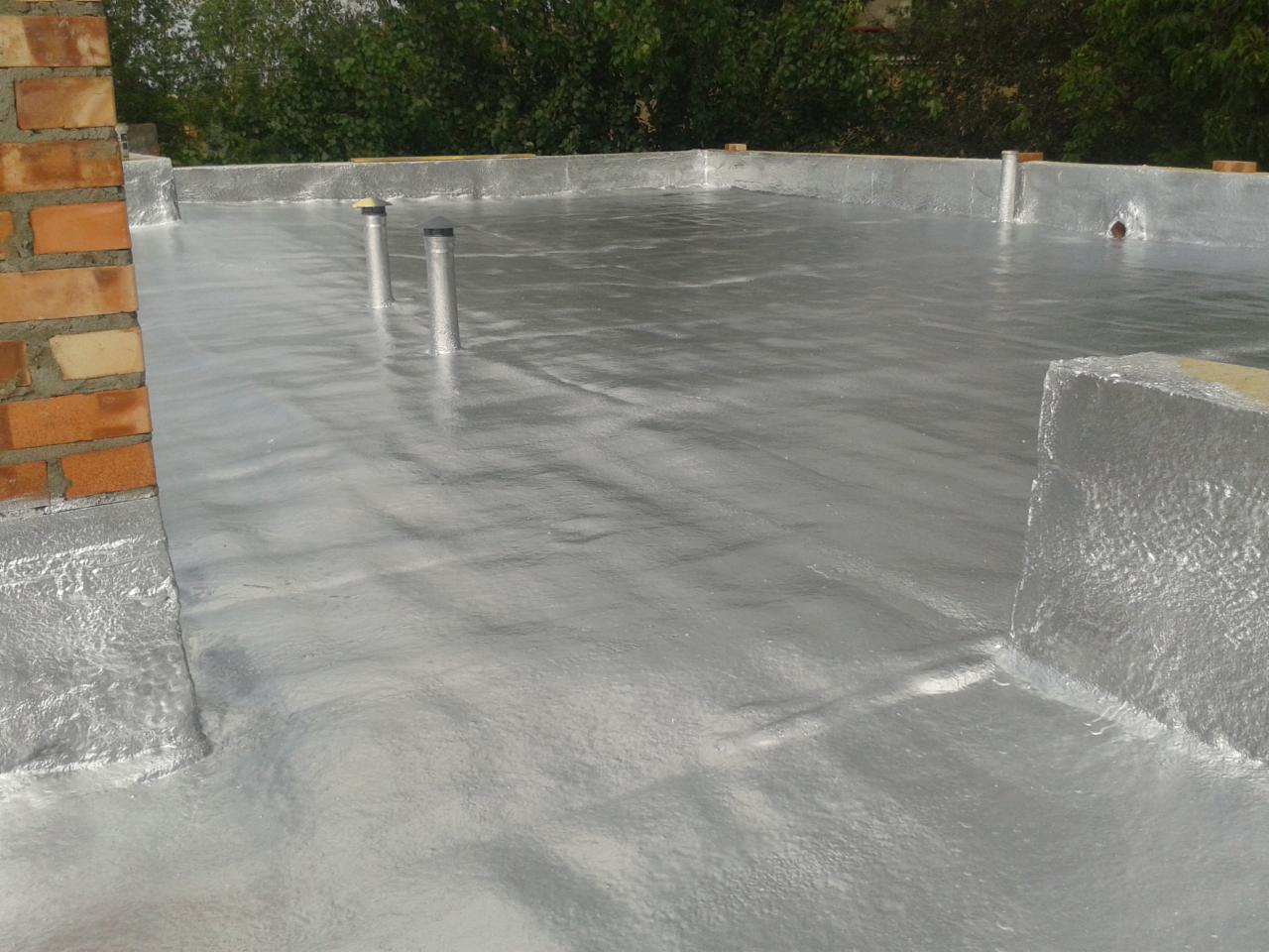 Szara, błyszcząca izolacja natryskowa na płaskim dachu z widocznymi kominkami wentylacyjnymi i fragmentem ceglanego komina.