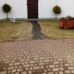 Biała ściana z brązową bramką, ścieżka z kamieni, dwa małe drzewka i drewniana donica. Ceglana kostka brukowa na pierwszym planie. Ujęcie w pochmurny dzień.