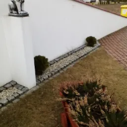 Biała elewacja domu z dekoracyjnym lwem na kolumnie, obok ścieżka z kamieni i rośliny w drewnianym wózku. Widok na dom z szarą dachówką w tle.