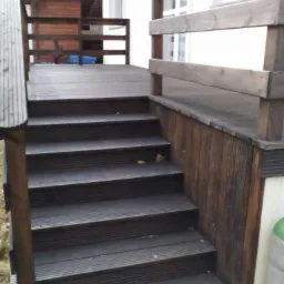 Drewniane schody prowadzące na taras z barierką, wykonane z ciemnego drewna. Widoczne detale konstrukcyjne i faktura drewna. Tło stanowi elewacja budynku.