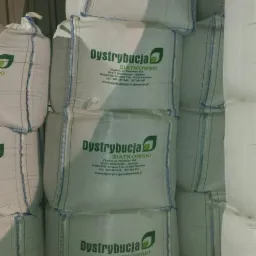 Stos białych worków z nadrukowanym logo firmy dystrybucyjnej z zielonym elementem graficznym w kształcie liścia, widoczne szwy z niebieską nicią.