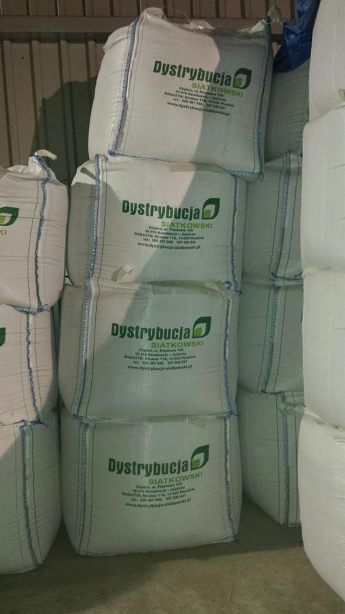 Stos białych worków z nadrukowanym logo firmy dystrybucyjnej z zielonym elementem graficznym w kształcie liścia, widoczne szwy z niebieską nicią.