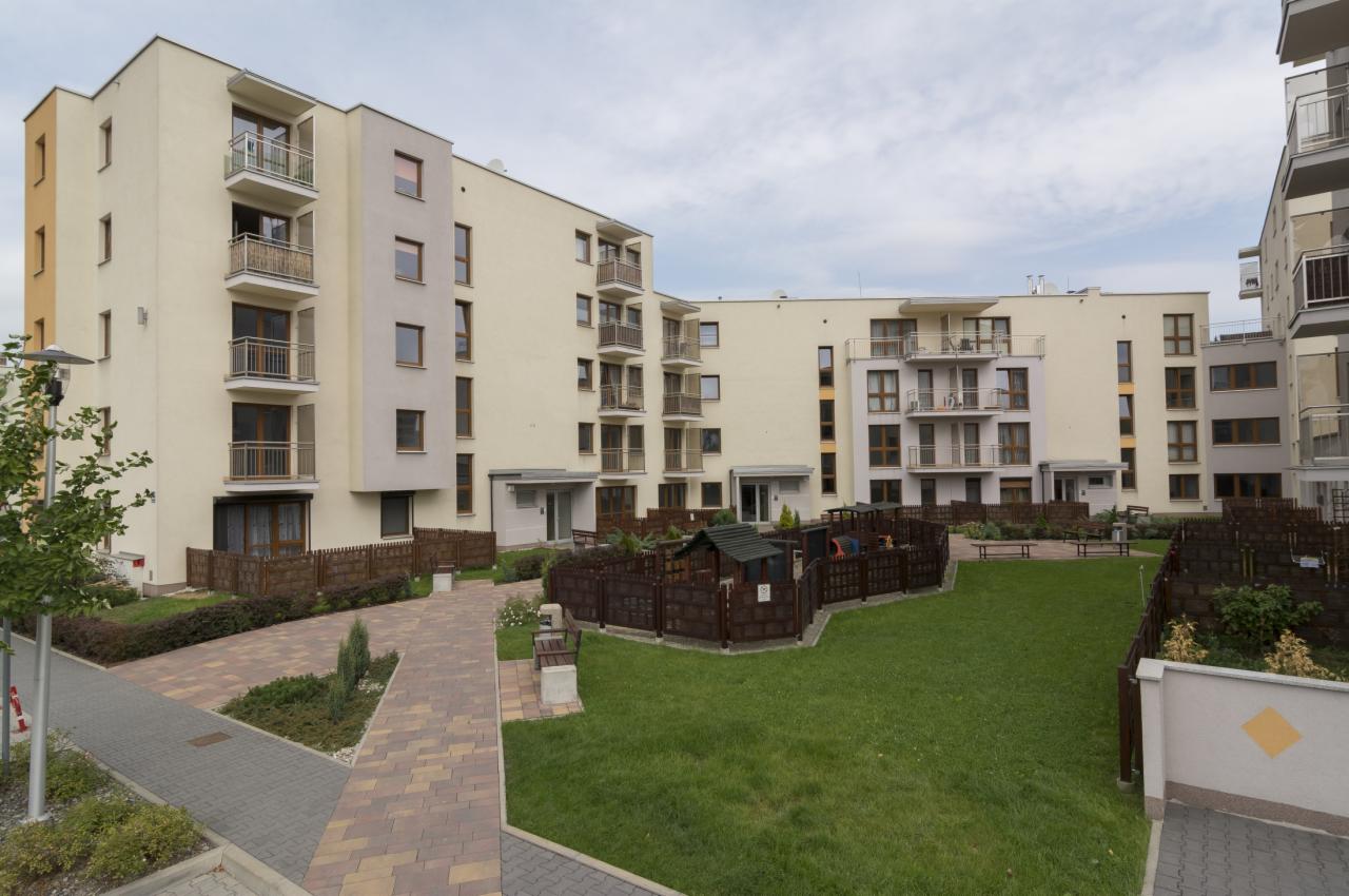 Nowoczesny kompleks apartamentowy z placem zabaw na dziedzińcu, zieleń trawnika i chodniki z kostki brukowej. Jasne elewacje budynków z balkonami.