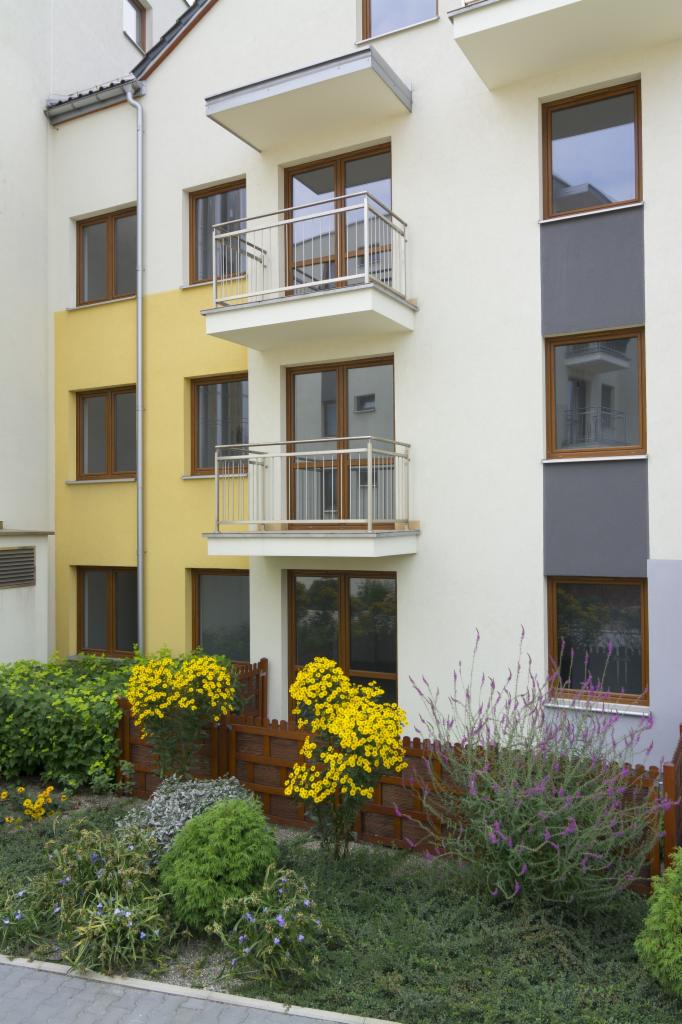 Elewacja budynku wielorodzinnego z dwoma balkonami z metalową balustradą. Żółte i szare akcenty kolorystyczne. Ogródek z niskim płotkiem i kwitnącymi krzewami.