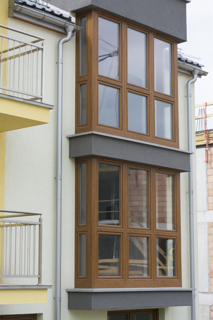 Dwukondygnacyjny wykusz z drewnianą stolarką okienną, widoczny z zewnątrz budynku z balkonem i rynnami. W tle mur z cegły i rusztowania.