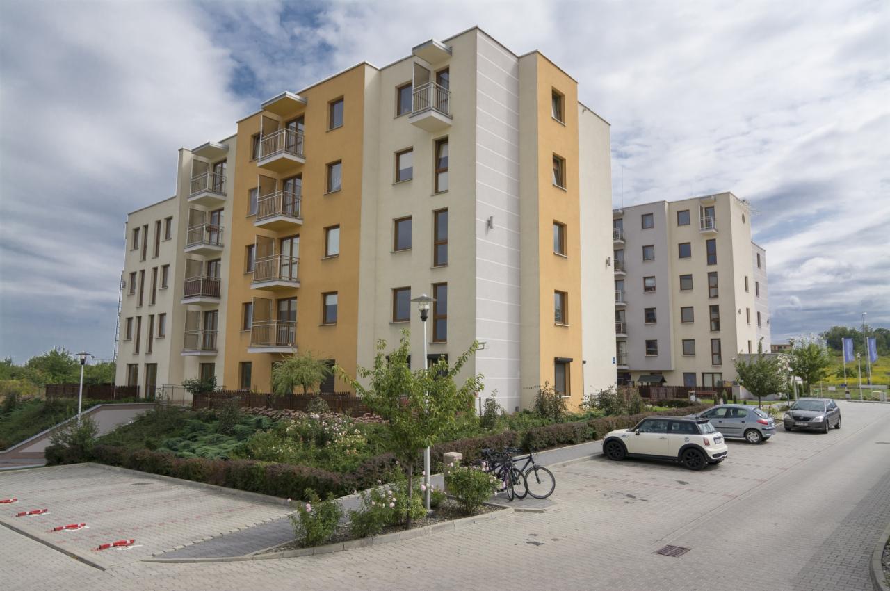 Nowoczesny budynek mieszkalny z balkonami, widok z poziomu parkingu z rowerami i zaparkowanymi autami. Budynek w odcieniach beżu i żółci.